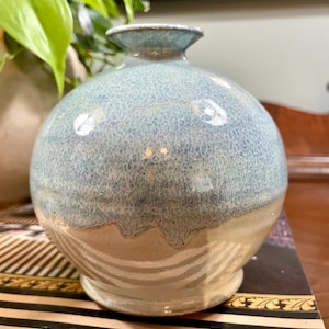 May include: A handcrafted ceramic vase with a rounded body and a narrow neck. The vase features a textured, speckled blue and white glaze on the upper half, transitioning to a beige base with horizontal stripes. A decorative piece for home decor.