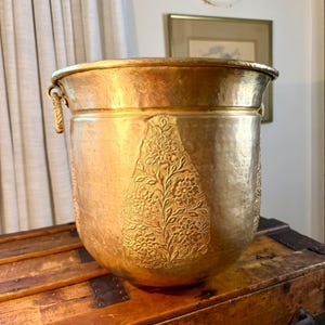 May include: A large, round, hammered brass bucket featuring a prominent embossed floral pattern on the front. The vessel has a wide, reinforced rim and small, decorative metal side handles with a rope-like texture.