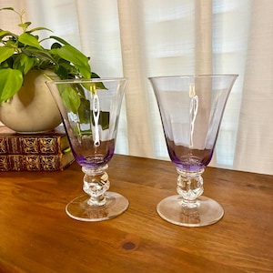 May include: Two clear wine glasses with purple bases and stems. The stems have a decorative, bulbous design. The glasses sit on a wooden surface, with a plant and books in the background.