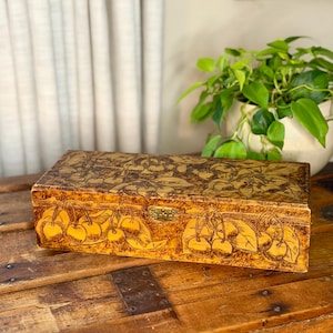 May include: A rectangular wooden box featuring a burnt-in wood design of cherry clusters with leaves on the lid and front panel. The box has a light brown, textured finish and a small, centered brass-colored metal latch on the front edge.