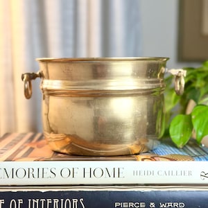 May include: A polished brass planter with a smooth, rounded body and two small ring handles on the sides. The vessel features a wide, open top and a slightly flared rim, showcasing a metallic gold finish with subtle signs of natural wear.