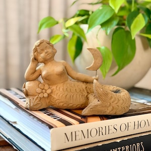 May include: A light tan ceramic figurine of a reclining mermaid with a sculpted flower at the base and a crescent moon shape held in one hand. The mermaid has detailed scale textures on the tail and rests on a stack of two hardcover books.