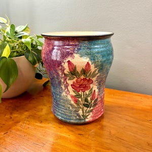 May include: A ceramic vase with a floral design. The vase features a painted rose with red petals and green leaves. The background has a blend of blue, purple, and cream colors. The vase is approximately 7 inches tall and 6 inches wide.