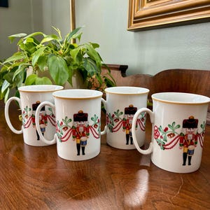 May include: Four white ceramic mugs with a gold rim and handle. Each mug features a colorful illustration of a nutcracker soldier in a red and gold uniform, surrounded by festive greenery and red ribbons. The mugs are arranged on a wooden surface.