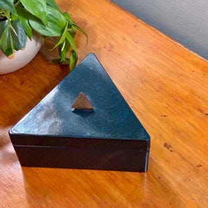 May include: A triangular, dark teal box with a copper-colored triangle on the lid. The box has a glossy finish and sits on a wooden surface. A potted plant is visible in the background.