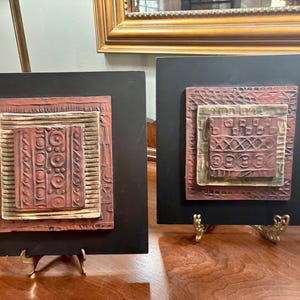 May include: Two decorative wall art pieces. Each features a square black background with a raised, textured, multi-layered design in shades of brown, tan, and green. The pieces are displayed on small, ornate gold stands.
