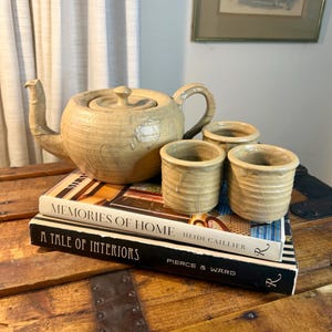 May include: A beige ceramic teapot with a matching lid and three small matching cups. The set features a textured, glossy glaze and is arranged on top of two stacked hardcover books titled Memories of Home and A Tale of Interiors.