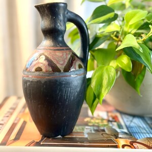 May include: A matte black ceramic pitcher with a single handle and a narrow neck. The body features a decorative band with abstract geometric patterns in shades of brown, cream, and muted red, set against a dark, textured, hand-finished surface.