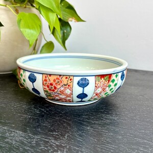 May include: A decorative ceramic bowl with a white interior and a blue rim. The exterior features alternating panels of floral patterns in red, orange, and green, and blue stylized flower designs. The bowl is on a dark surface.