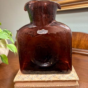 May include: A large, square-shaped, dark purple Blenko glass vase with a wide, flared opening. The vase has a textured surface and a circular indentation on the front. It sits atop two antique books. The Blenko Handcraft label is visible.