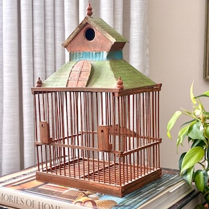 May include: A wooden birdcage with a brown slatted base and a multi-tiered roof. The roof features a green painted finish, a small arched wooden door, and a circular opening at the top. The structure includes vertical brown bars and two small wooden brackets.