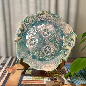 May include: A round ceramic plate with a scalloped edge, featuring a glossy teal and white glaze. The surface is embossed with intricate marine life patterns, including sea turtles, shells, and three prominent raised starfish in the center.