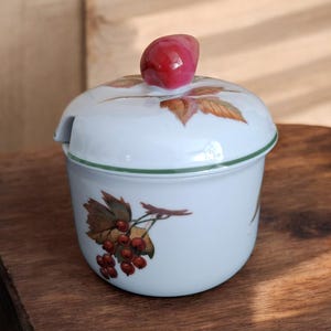Può includere: Zuccheriera in ceramica bianca con coperchio e manico a forma di frutto rosso. La ciotola presenta un bordo verde ed è decorata con illustrazioni di bacche rosse e foglie. Una piccola apertura laterale è per un cucchiaio.