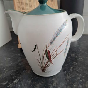Può includere: Teiera in ceramica bianca con coperchio e manico verde acqua. La teiera è decorata con un motivo floreale nei toni del marrone, verde e blu. Un articolo di cucina.