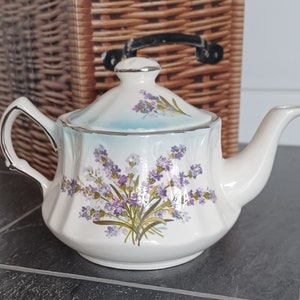 Könnte beinhalten: Eine weiße Teekanne mit einem Lavendel-Blumenmuster. Die Teekanne hat einen Deckel und einen Ausguss. Die Teekanne steht auf einer braunen Weidefläche.