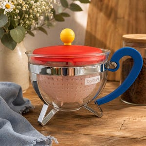 Op de afbeelding: Een theepot van helder glas met een rood deksel, een gele knop en een blauwe handgreep. De theepot heeft een verchroomd metalen frame met drie pootjes en een lichtroze zeef aan de binnenkant. De merknaam bodum staat in wit op het glas.