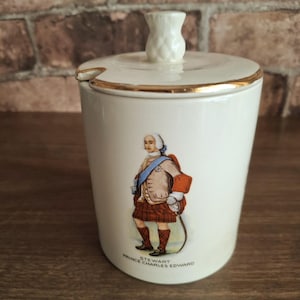 May include: A cream-colored ceramic jar with a lid featuring a thistle-shaped handle and a gold-trimmed rim. The front displays a colorful illustration of Prince Charles Edward Stewart in traditional Scottish attire, with his name printed below.