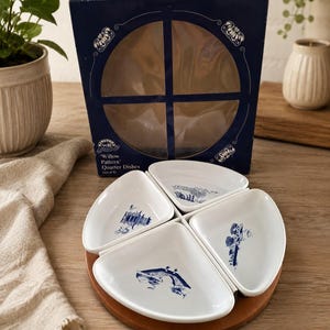 May include: A set of four white ceramic quarter dishes with blue willow pattern illustrations, arranged on a round wooden tray. The dishes are presented in a dark blue box labeled Willow Pattern Quarter Dishes, featuring a circular cutout window.