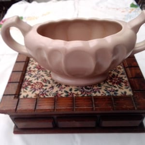 May include: A pale pink ceramic planter with two handles sits on a wooden box with three drawers. The box is lined with a floral fabric.