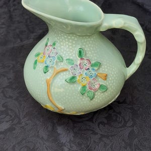 May include: A light mint green ceramic jug featuring a textured dotted surface and a raised relief design of branches with small colourful flowers in shades of pink, blue, and yellow. The jug has a curved handle and a wide pouring spout.