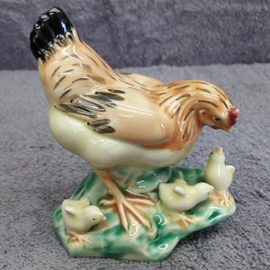 May include: A ceramic figurine of a brown and white hen with black markings on its head, standing on a green base with three smaller chicks. The hen is looking down at the chicks.