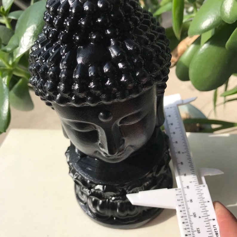 Puede incluir: Estatua de cabeza de Buda negra con un mo&ntilde;o detallado y una expresi&oacute;n serena. La estatua mide aproximadamente 18 cm de alto y est&aacute; hecha de un material negro liso.