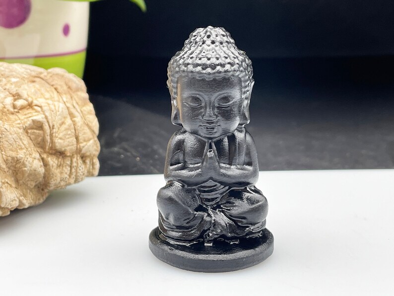 May include: A small black Buddha statue sitting in a meditative pose with hands clasped in prayer. The statue is made of a smooth, polished material and has a serene expression.