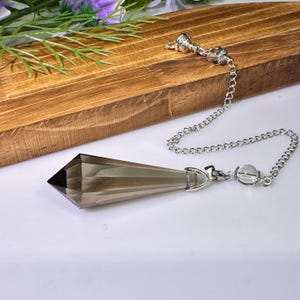 May include: A faceted, smoky-colored crystal pendulum with a pointed tip, attached to a silver-toned metal chain. The chain features a small, clear spherical bead near the crystal attachment point and a decorative silver-toned end piece.