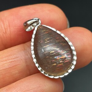 May include: A teardrop-shaped pendant with a silver setting and a iridescent sunstone gemstone. The stone has a brown base with streaks of rainbow colors.