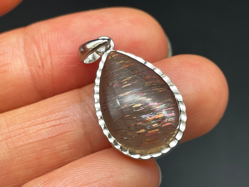 May include: A teardrop-shaped pendant with a silver setting and a iridescent sunstone gemstone. The stone has a brown base with streaks of rainbow colors.