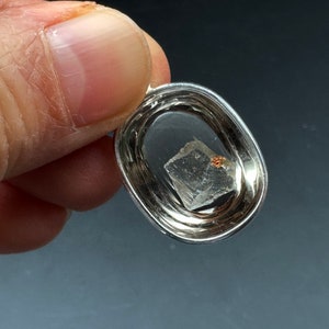 May include: A silver pendant with a clear crystal inside. The crystal is a cube shape with a small, reddish-brown inclusion.
