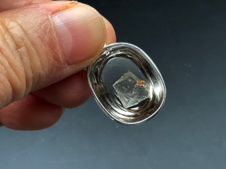 May include: A silver pendant with a clear crystal inside. The crystal is a cube shape with a small, reddish-brown inclusion.