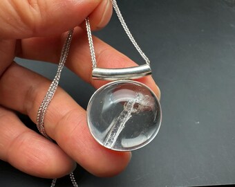 May include: A clear quartz pendant necklace with a silver chain. The pendant is a round, transparent stone with a visible inclusion. The necklace features a silver bar above the pendant, connecting to a delicate chain.