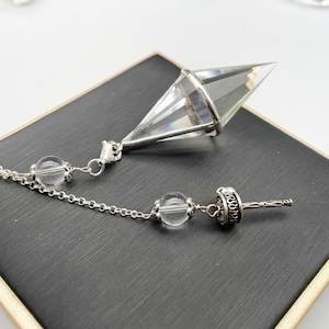 May include: A clear quartz crystal pendulum with a silver chain and beads. The pendulum is in the shape of a pyramid and has a silver tip.
