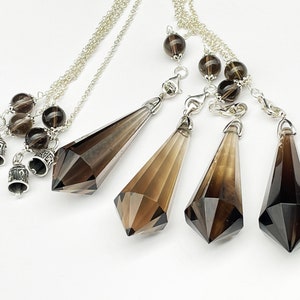 Könnte beinhalten: Vier rauchquarzkristall-Pendel mit silbernen Ketten und Perlen. Die Pendel hängen an einer weißen Oberfläche.