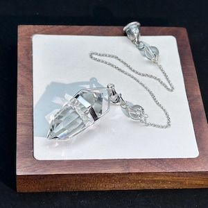 May include: A clear, faceted, pointed crystal pendulum suspended from a delicate silver-toned metal chain. The chain features two small, round, clear crystal beads and a silver-toned metal end cap, creating a balanced, dangling design.