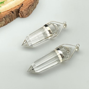 Puede incluir: Dos colgantes de punta de cristal de cuarzo transparente con engastes de plata. Los colgantes cuelgan de un fondo blanco.
