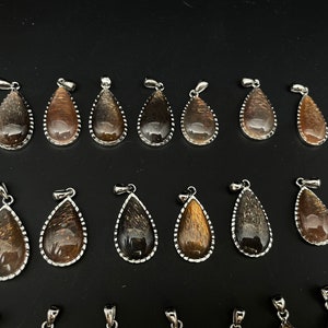 May include: A collection of 18 gold sunstone pendants in a teardrop shape with silver settings. The pendants are arranged in two rows on a black surface.