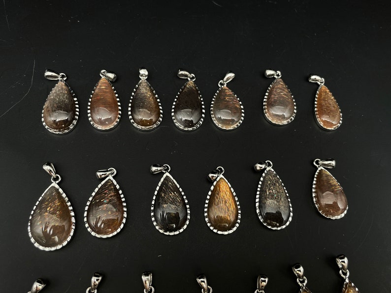 May include: A collection of 18 gold sunstone pendants in a teardrop shape with silver settings. The pendants are arranged in two rows on a black surface.