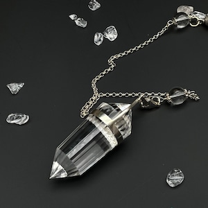 Op de afbeelding: Een heldere, gefacetteerde, zeshoekige kristallen hanger bevestigd aan een zilverkleurige metalen ketting. Het kristal heeft een puntige vorm en een platte bovenkant die wordt vastgehouden door een metalen dop. De ketting bevat een kleine, heldere kraal en een decoratief eindstuk.