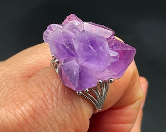 Puede incluir: Un anillo de plata con un gran racimo de cristales de amatista. El cristal es de color morado oscuro y tiene una textura natural y rugosa.