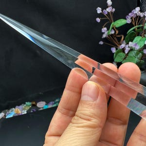 Könnte beinhalten: Ein klarer, spitzer Kristallstab mit facettierter Basis. Der Stab wird vor einem dunklen Hintergrund gehalten, mit einem kleinen Blumenarrangement im Hintergrund. Der Kristall ist etwa 18 cm lang.
