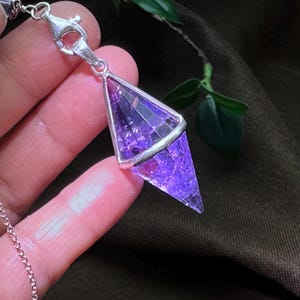 May include: A faceted, double-terminated purple crystal pendant encased in a silver-toned metal frame. The crystal has a translucent, textured interior with deep violet hues. It is attached to a silver-toned lobster clasp and a thin metal chain.