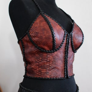 Peut inclure: Un haut en cuir marron avec un motif peau de serpent et des bordures tressées noires. Le haut a un décolleté en V et une fermeture éclair à l'avant.