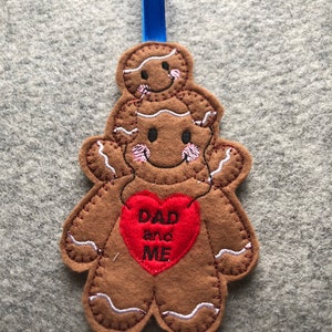 May include: A brown felt gingerbread man ornament with a red heart that says "DAD and ME" on it. The gingerbread man is holding a smaller gingerbread man on top of its head.