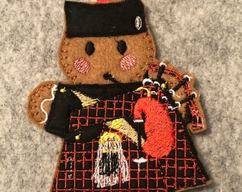 May include: A gingerbread man ornament wearing a black hat and a red and black plaid kilt. He is playing a bagpipe with a red bag and black and white pipes.