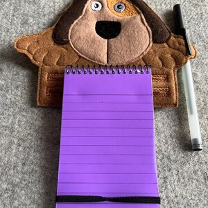 May include: A brown felt dog-shaped notepad holder with a purple lined notepad and a black pen. The notepad has a black elastic band to hold it closed.