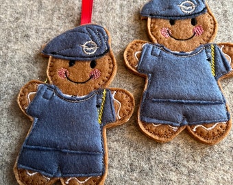 May include: Two felt gingerbread ornaments, each wearing a blue uniform with a yellow stripe and a blue hat with a white logo. The ornaments are hanging from red ribbons.