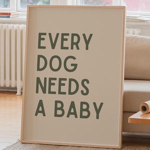 Op de afbeelding: Een ingelijste print met de tekst "EVERY DOG NEEDS A BABY" in groene letters. De print staat in een lichtgekleurde houten lijst en leunt tegen een muur in een woonkamer. Links staat een grote plant in een witte pot.