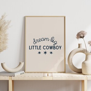 May include: A framed print with a beige background and the text "dream big LITTLE COWBOY" in dark blue with three stars underneath. The print is sitting on a light wood shelf with two white ceramic vases and dried pampas grass.